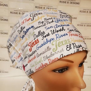 May include: White surgical scrub hat with a colorful print of Texas cities and landmarks. The hat has a tie on the back and a small triangle pattern on the tie.