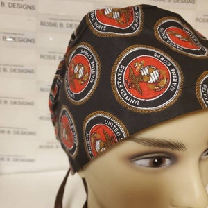 May include: A black scrub hat with a repeating pattern of the United States Marine Corps emblem. The emblem is red, gold, and white with a rope border. The hat has brown ties.