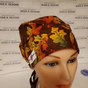 May include: A brown surgical scrub hat with a fall leaf print. The hat has a brown tie and a pink heart with the text "Rosie Designs" on it.