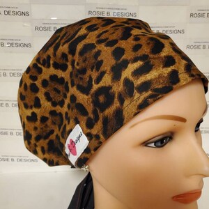 May include: A brown and black leopard print surgical scrub hat with a black tie. The hat has a white label with a pink heart and the text "Rosie B. Designs".