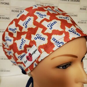 May include: A white and red scrub hat with a blue and white outline of the state of Texas repeated across the fabric. The hat has a small label that says "Rosie B. Designs" with a pink heart.