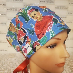 May include: A blue surgical scrub hat with a colorful print featuring Frida Kahlo, parrots, and flowers. The hat has a red tie and a small label that says "B. Designs".