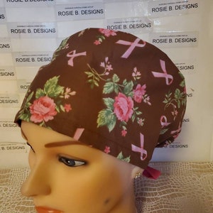 May include: A brown surgical scrub hat with a pink floral pattern and pink ribbon accents. The hat is made by Rosie B. Designs.