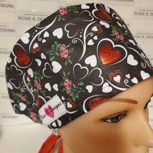May include: A black scrub hat with a red and white heart pattern. The hat has a red tie and a small label that says "Rosie B. Designs".