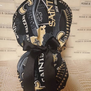 May include: A black and gold fabric scrub hat with a black bow. The hat features the New Orleans Saints logo and the words "Saints" and "New Orleans".