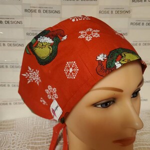 Peut inclure: Un bonnet de travail rouge à thème de Noël avec un personnage vert du Grinch et des flocons de neige blancs. Le bonnet a une cravate à l'arrière.