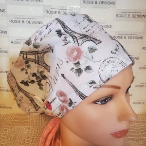 May include: A white surgical scrub hat with a pink and gray Eiffel Tower design. The hat has a tie on the back and a button on the top. The hat is made of a soft, comfortable fabric.