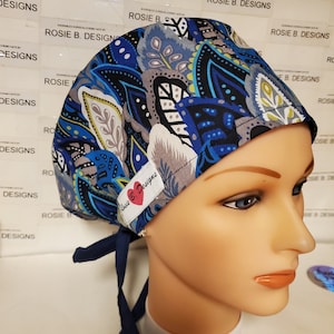 Könnte beinhalten: Eine blaue, graue und schwarze gemusterte OP-Mütze mit einem roten Herzlogo, auf dem "Rosie B. Designs" steht.