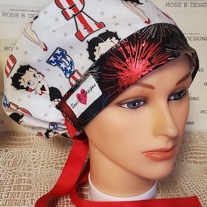 Könnte beinhalten: Eine weiße OP-Haube mit einem rot-weiß-blauen amerikanischen Flaggenmuster und einem rot-weiß-blauen Feuerwerksmuster. Die Haube hat eine rote Schleife und zeigt ein Cartoon-Bild von Betty Boop. Die Haube ist mit "Rosie B Designs" beschriftet.