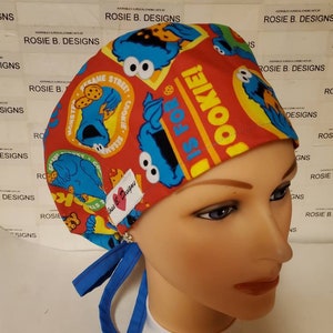 May include: A red, blue, and yellow surgical scrub hat with a Sesame Street theme. The hat features Cookie Monster and the text "It's for Cookie!" The hat has blue ties.