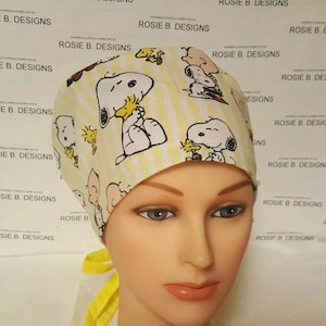 May include: A yellow and white striped surgical cap featuring Snoopy and Woodstock characters. The cap has a tie in the back and the text "ROSIE B. DESIGNS" is printed on the background.