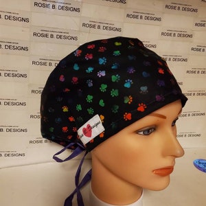 Op de afbeelding: Een zwarte chirurgische scrub-muts met een kleurrijk pootprintpatroon. De muts heeft een paarse stropdas en een wit label met de tekst "Rosie B. Designs" in rood.