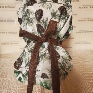 May include: A white scrub hat with a brown ribbon and a pine tree print. The hat has a brown ribbon tied in a bow at the top. The print features pine trees, pine cones, and swirls.