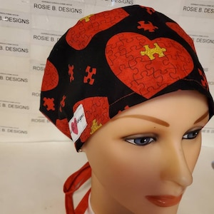 May include: A black scrub hat with a red and gold puzzle piece heart design. The hat has a red tie and the words "Rosie B. Designs" are printed on the hat.