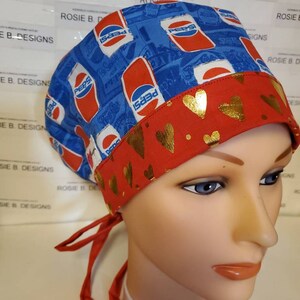 May include: A blue surgical scrub cap featuring a Pepsi logo design and a red band adorned with gold hearts. It has red ties for fastening. The cap is displayed on a mannequin head.