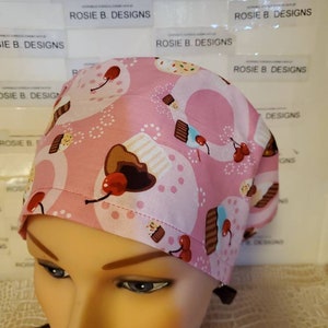 May include: A pink surgical scrub cap with a cupcake pattern. The cap features white frosting, chocolate cupcakes, and red cherries. The text "ROSIE B. DESIGNS" is visible on the background.