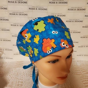 May include: A blue surgical scrub hat with a colorful pattern of puzzle pieces and cartoon characters. The hat has a tie on the back.