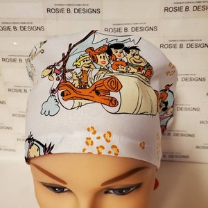 May include: White surgical scrub hat with a cartoon image of the Flintstones family riding in their car. The hat has a leopard print pattern on the top.