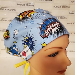 May include: Blue and yellow surgical scrub hat with a comic book design featuring Sonic the Hedgehog. The hat has a yellow tie and a pink heart with the text "Rosie B. Designs".