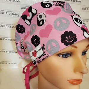May include: Pink surgical cap with a black and white peace sign, yin yang, and smiley face pattern. The cap has a pink tie and the text "Love is Blind!" with a heart and the word "Ananda" in a smaller font.
