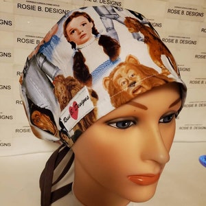 May include: A patterned surgical cap featuring characters from The Wizard of Oz, including Dorothy and the Cowardly Lion. The cap is white with a brown tie and a small label that reads "Rosie B. Designs".