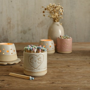 Puede incluir: Un portavelas de cerámica blanca con un diseño de corazón y las iniciales "L + S" grabadas en la parte delantera. El portavelas está lleno de cerillas de colores y está sentado en una mesa de madera junto a otros portavelas de cerámica.