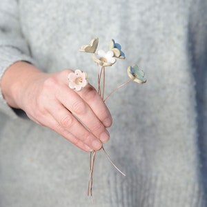 May include: A small bouquet of four handcrafted ceramic flowers attached to thin copper wire stems. The flowers feature five petals each in soft pastel shades of pale pink, white, and light blue, with a small circular center in each blossom.