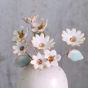 Op de afbeelding: Een keramische vaas bevat een boeket witte bloemen met gouden accenten. De bloemen hebben een madeliefachtige vorm en zijn gerangschikt met koperkleurige draadstelen en lichtgroene bladeren. De vaas is een gespikkelde gebroken witte kleur.