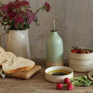 Può includere: Una scena di cucina rustica con una bottiglia di olio verde chiaro, ciotole in ceramica e un vaso con fiori rosa. Pane a fette poggia su un tagliere di legno, insieme a pomodori, ravanelli e verdure. La scena è ambientata su un tavolo di legno.