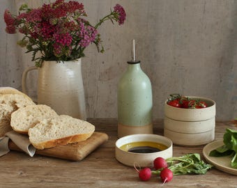 May include: A rustic kitchen scene featuring a light green oil bottle, ceramic bowls, and a vase with pink flowers. Sliced bread rests on a wooden cutting board, alongside tomatoes, radishes, and greens. The scene is set on a wooden table.