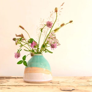 Könnte beinhalten: Eine Keramikvase mit türkisfarbenem Oberteil, beigem Streifen und weißem Sockel, gefüllt mit Wildblumen. Die Blumen beinhalten rosa Klee und Gräser. Die Vase steht auf einer verwitterten, hellen Holzoberfläche.