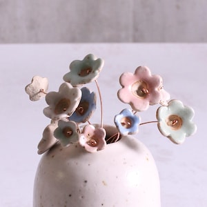 May include: A white ceramic vase with a speckled glaze holds a bouquet of pastel ceramic flowers. The flowers are in shades of pink, blue, and green, and each has a small copper button in the center.