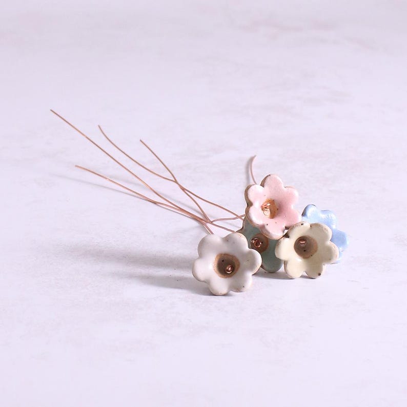 May include: A cluster of small, five-petaled ceramic flowers in pastel shades of pink, blue, and cream. Each individual flower is attached to a thin, flexible copper-colored wire stem, creating a delicate floral arrangement with a smooth finish.