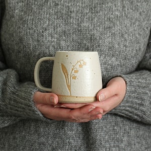 Puede incluir: Una taza de cerámica color crema con asa, con un diseño floral. La taza tiene una textura moteada y una base marrón claro. El diseño floral es un lirio de los valles estilizado en un color dorado cálido. La taza se sostiene con ambas manos.