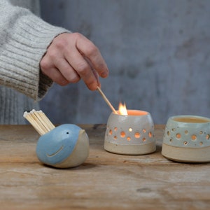 May include: A ceramic bird match holder with a blue body and a beige beak, holding a stack of matches.  Two ceramic candle holders with a white glaze and beige base, one with a lit candle inside.