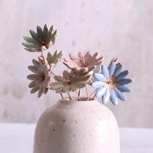 Puede incluir: Un pequeño jarrón de cerámica blanca con un esmalte moteado contiene un ramo de flores de cerámica en colores pastel. Las flores son en tonos de azul, rosa y verde.