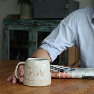 Può includere: Tazza in ceramica maculata con la scritta "Daddy". La tazza è bianco sporco con base e manico beige. La tazza è appoggiata su un tavolo di legno.