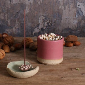May include: A pink ceramic match holder filled with white-tipped matches, next to a light green incense holder with a pink incense stick. The items are on a wooden surface with nuts scattered around.