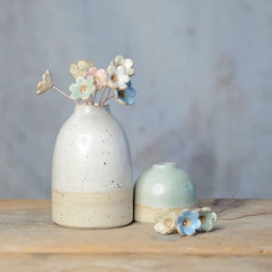 May include: A tall white speckled ceramic vase and a shorter light green ceramic vase. The tall vase holds several small ceramic flowers on thin copper wires in shades of blue, pink, and white. Three additional ceramic flowers lie nearby.
