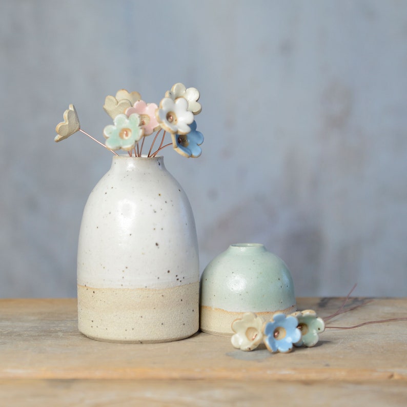 May include: A tall white speckled ceramic vase and a shorter light green ceramic vase. The tall vase holds several small ceramic flowers on thin copper wires in shades of blue, pink, and white. Three additional ceramic flowers lie nearby.