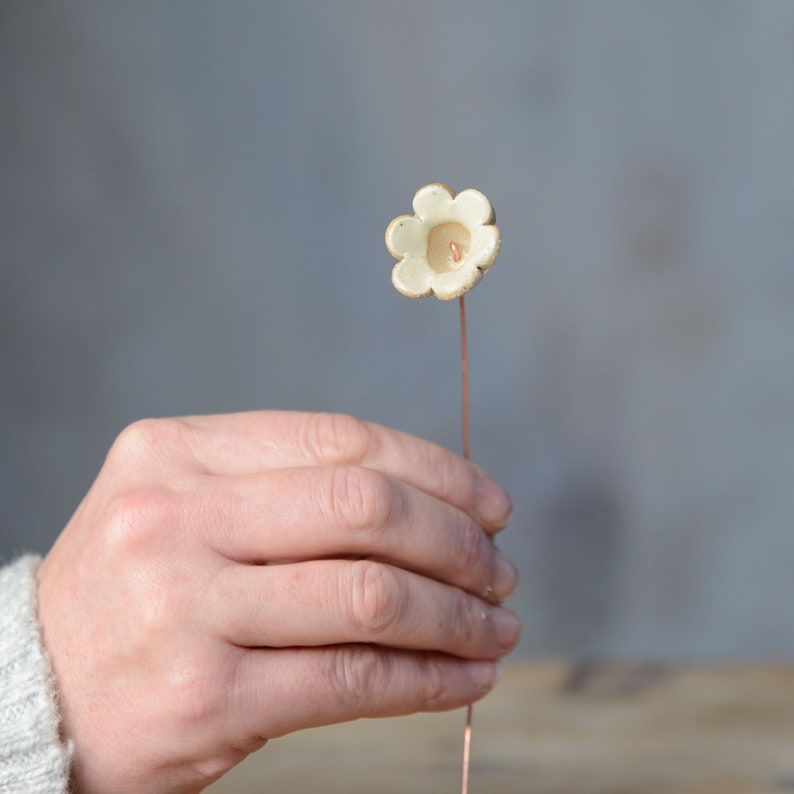May include: A small, off-white ceramic flower ornament with six rounded petals and a central indentation, attached to a thin, straight copper-colored metal wire stem. The ceramic surface has a matte, slightly textured appearance.