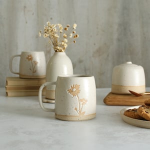 Op de afbeelding: Een verzameling handgemaakte keramische items, waaronder mokken, een vaas met gedroogde bloemen en een pot met deksel. De mokken hebben een madeliefjesontwerp. De items zijn in neutrale tinten met een gespikkelde afwerking.