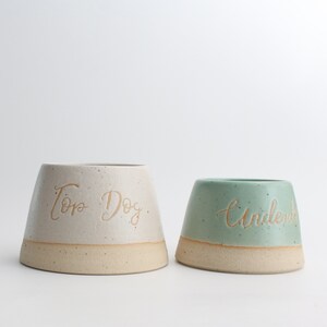 May include: Two ceramic dog bowls, one white with the text "Top Dog" and one green with the text "Underdog".
