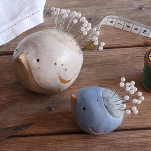 May include: Two ceramic bird-shaped pincushions, one larger and off-white, the other smaller and light blue, both filled with white-topped sewing pins. A white measuring tape, a spool of green thread, and metal scissors are positioned nearby.