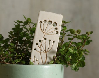 Puede incluir: Marcaplanas de cerámica blanca con un diseño recortado de dos ramitas de flores. El marcador está en una maceta verde con una pequeña planta verde.