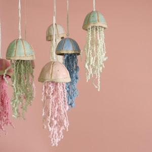 Peut inclure: Ornements de méduses en céramique faits à la main, aux couleurs pastel variées : rose, bleu et vert. Chaque méduse est ornée d'une base en fil texturé, suspendue par une ficelle blanche. Le fond est rose uni.