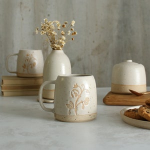 May include: Three white ceramic mugs with a floral design in a light brown color. The mugs are on a white surface with a vase of dried flowers and a wooden cutting board with cookies.