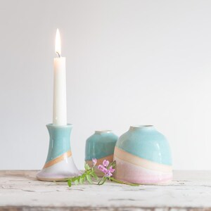May include: A lit white candle in a turquoise and gold ceramic candlestick holder, with two matching turquoise and pink vases. The candle and vases are on a white wooden surface. A sprig of purple flowers is in front of the vases.