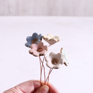May include: A collection of five small, hand-sculpted ceramic flowers in pastel shades of blue, pink, cream, and white. Each flower features a central indentation and is attached to a thin, flexible copper-colored wire stem for display.