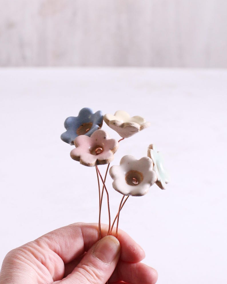 May include: A collection of five small, hand-sculpted ceramic flowers in pastel shades of blue, pink, cream, and white. Each flower features a central indentation and is attached to a thin, flexible copper-colored wire stem for display.